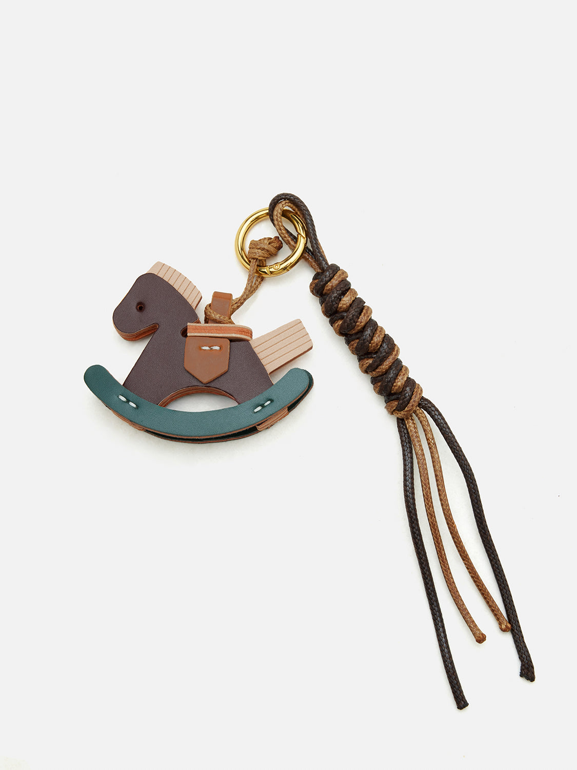 A small leather keychain charm shaped like a rocking horse, with a dark brown body, tan saddle and mane, a muted teal curved base, a gold-tone key ring, and a knotted braided leather tassel on a light gray background.
